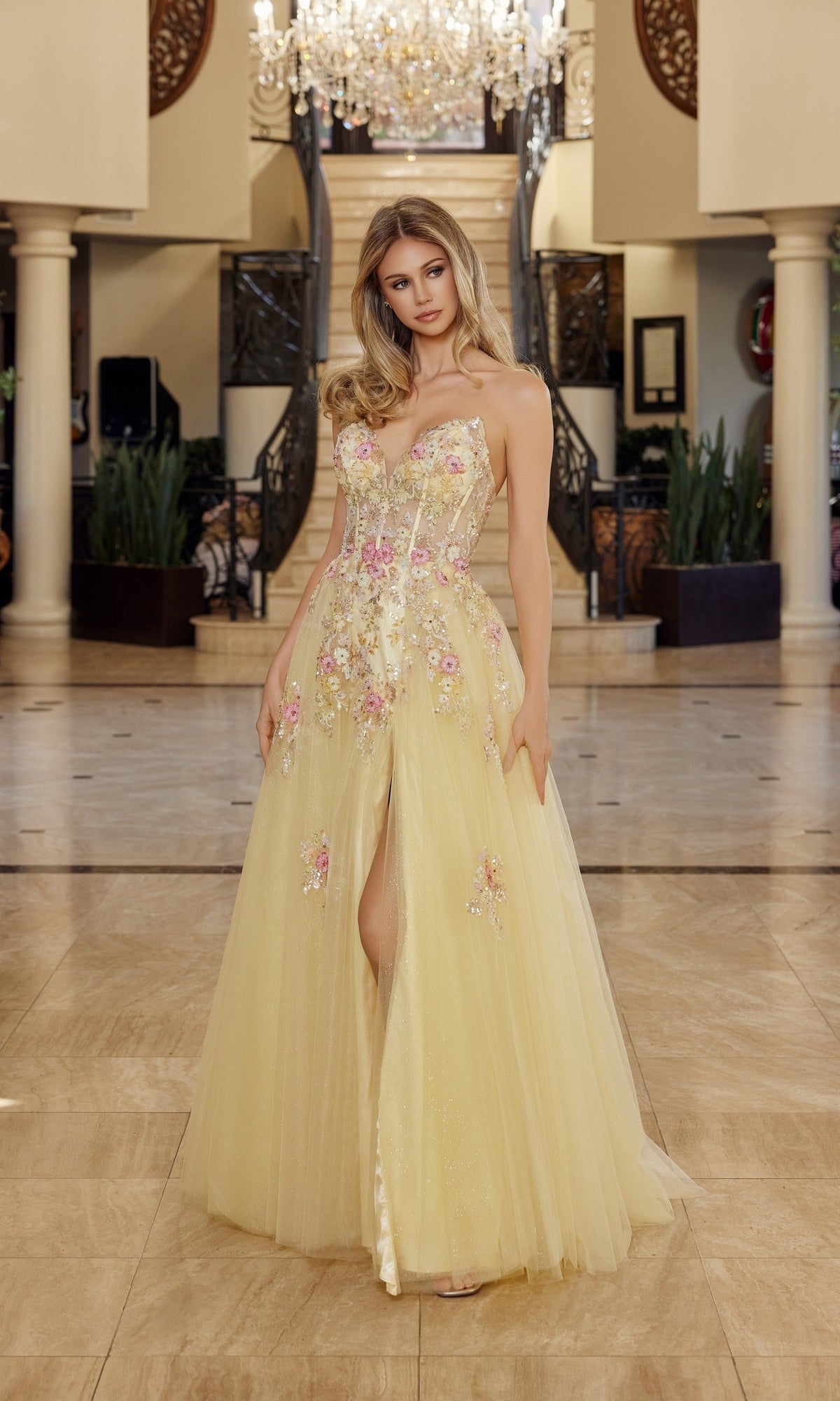 A woman stands indoors wearing the Nox Anabel Q1773 long yellow prom dress with a strapless corset bodice, 3D floral appliques, and a front slit. A staircase and chandelier can be seen in the background.
